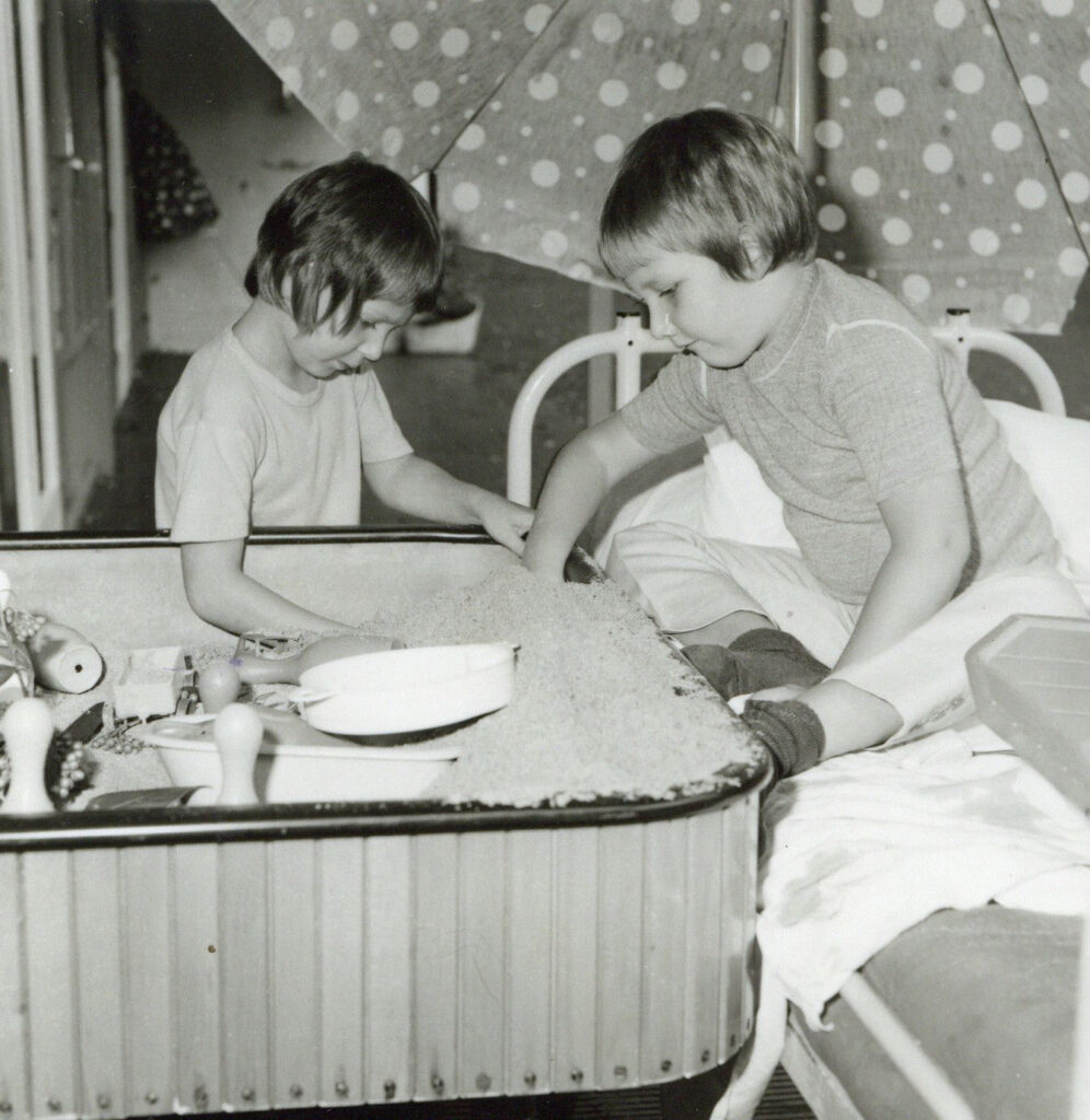  Twee patiëntjes van de Hemofiliekliniek met mobiele zandbak bij het bed van een van de patiëntjes